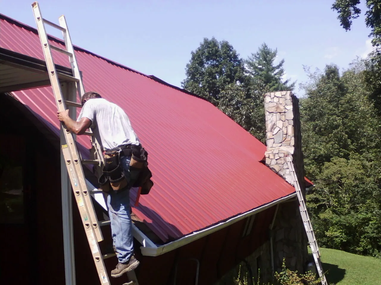 Roofer on a ladder beside a red metal roof and stone chimney — Soffit & Fascia Repair by Roofing Brothers in Marion Center